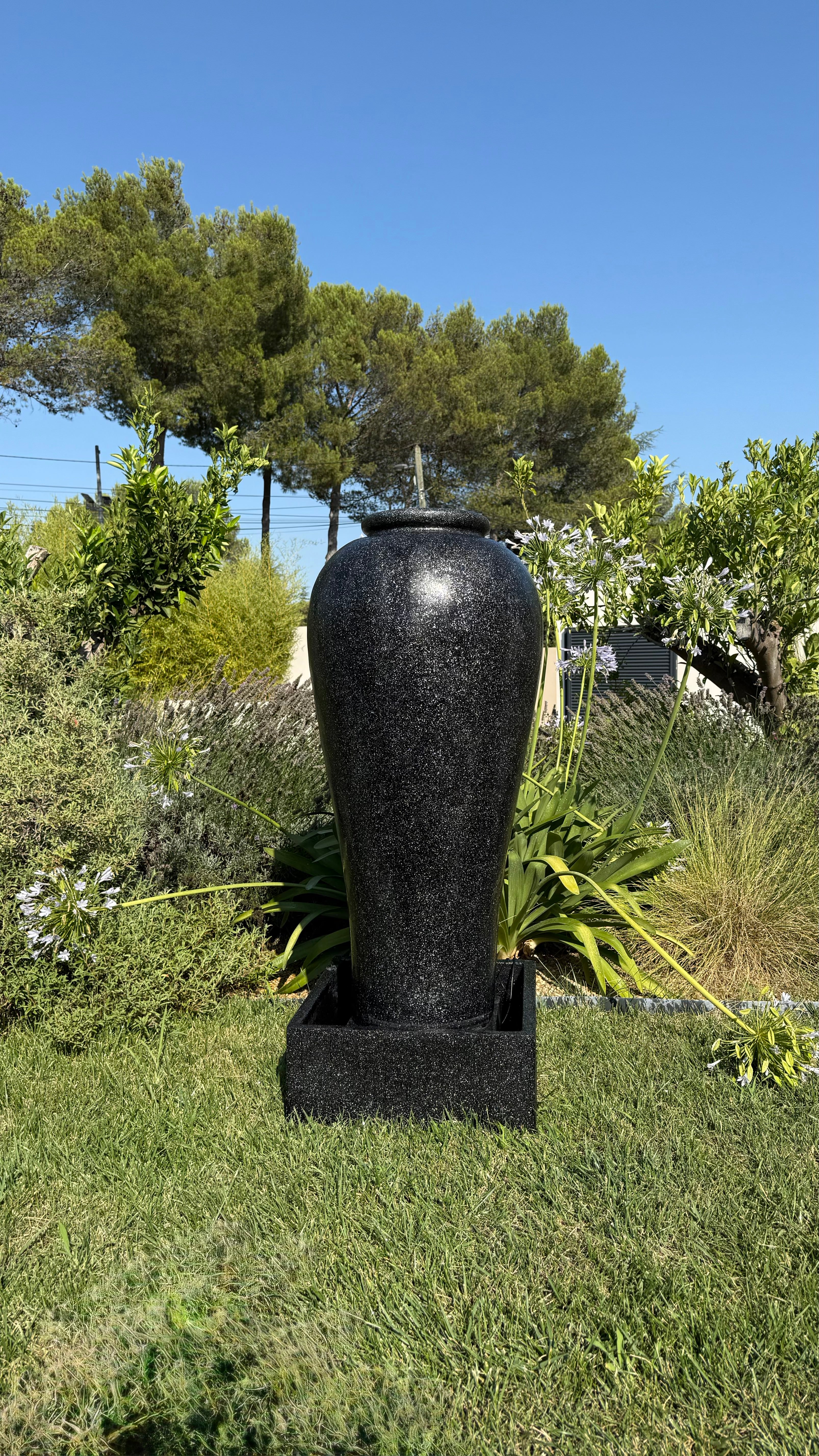 Fontaine extérieure noire de style moderne, modèle grand vase de l'Ensemble Socle Noir Moderne, installée dans un jardin verdoyant.