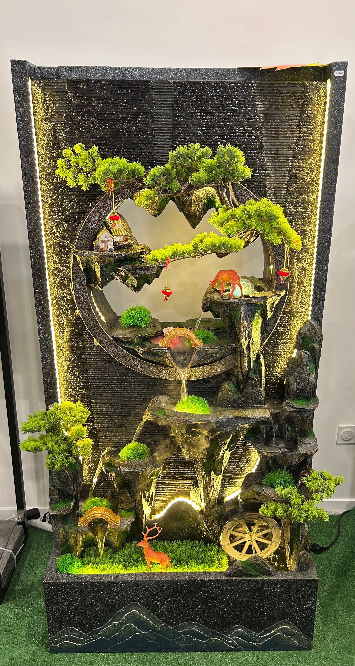 Fontaine Asiatique Dongguan éclairée en intérieur, avec détails de cerfs, maison miniature et chute d’eau, créant une atmosphère apaisante.